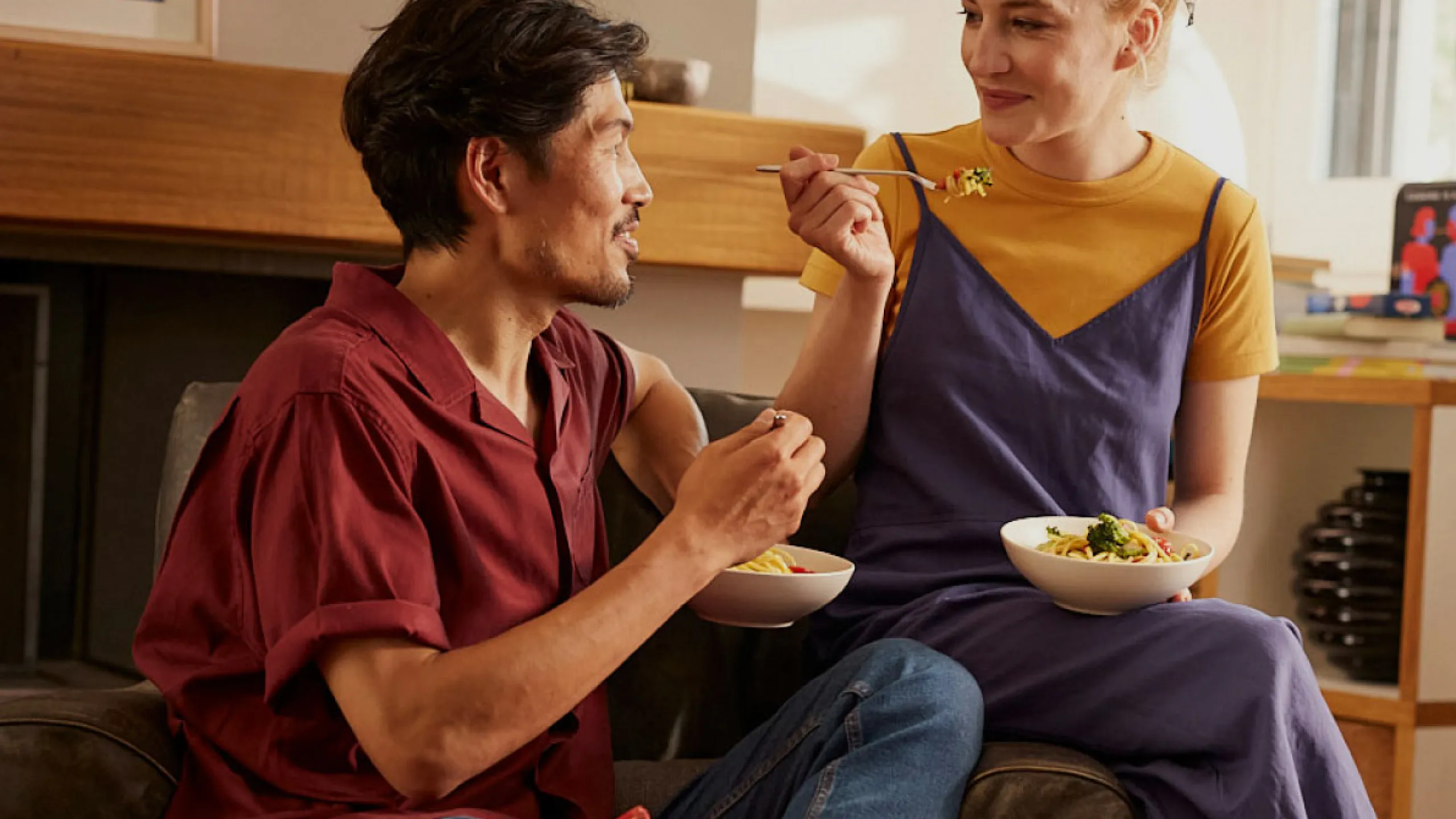 Young couple sharing a meal. Each is eating from a bowl of pasta. The man wears a red shirt, the woman wears a yellow shirt and blue dungarees. | GEEU-38i2gKIv1LRQ1NxTXQoXeR